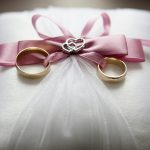 Close-up of elegant wedding rings on a silk pillow with a pink ribbon and a heart decoration, symbolizing love.